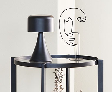 Minimalistisk sort lampe og ansiktsformet trådskulptur på et lite glassbord