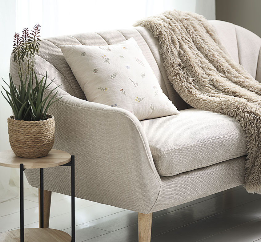 RASMUS kunstig plante i STEFAN potte, AGERTIDSEL pute og MYRFIOL langhåret pledd Kunstig aloe vera plante med blomster, brodert pute og langhåretpledd på en beige sofa