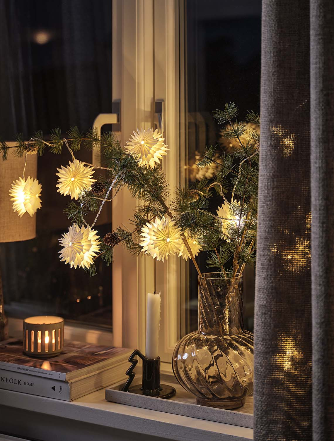 Stjerneformede lyslenker arrangert på furugrener i en vase som lyser opp en vinduskarm om natten