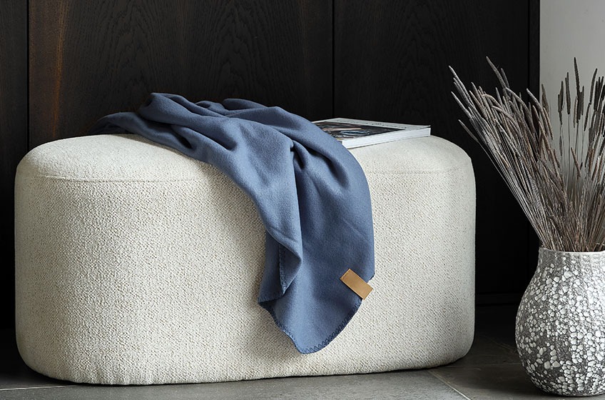 Off-white pouffe with a blue throw on top beside a grey and white vase 