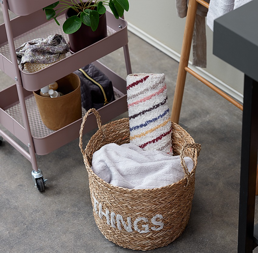 Bathroom accessories on a trolley next to a wicker storage basket with towels and a bath mat in it 