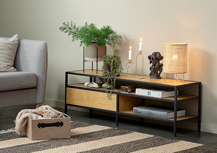 Et hjørne av en stue med en sofa, en kurv, en liten bordlampe og et moderne TV-bord