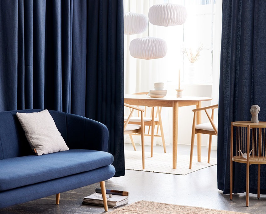 AMUNGEN gardin bak GISTRUP sofa og EGELEV sodebord. I bakgrunnen står MARSTRAND spisebord, ARNBORG spisestoler og VALDER lamper i bakgrunnen Blå gardin bak en sofa skiller en stue fra en spisestue