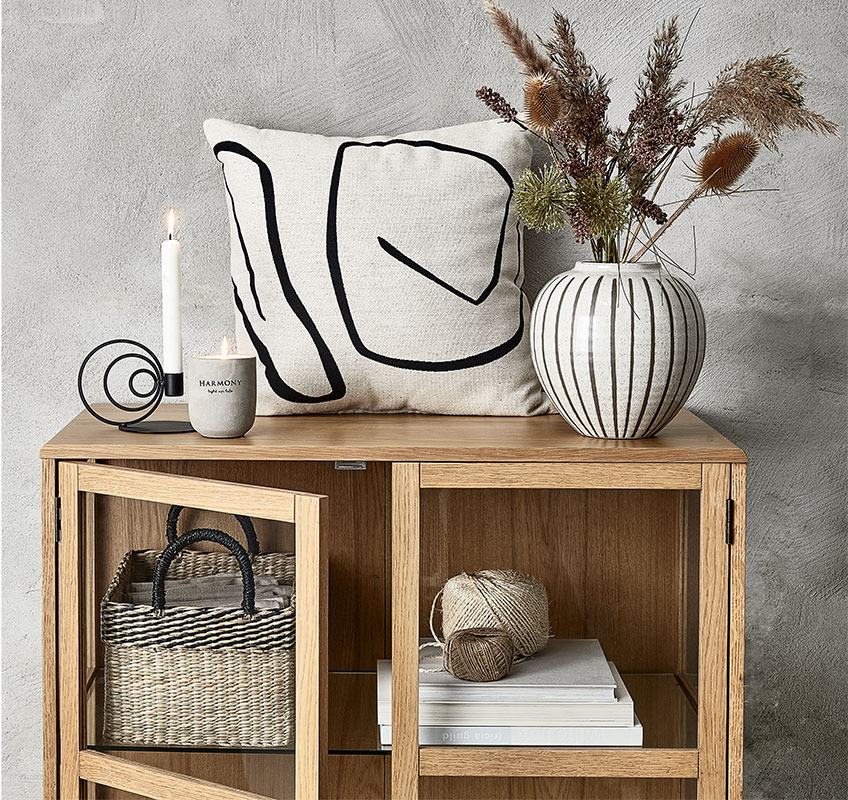 SOFUS vase, KEVIN lysestake, BOSSE stearinlys, og HVITVEIS pyntepute Et vitrineskap med bøker, kurv, lys, pyntepute og vase