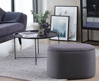 En rund puff i grå i en stue med et hjørne bord og en sofa
